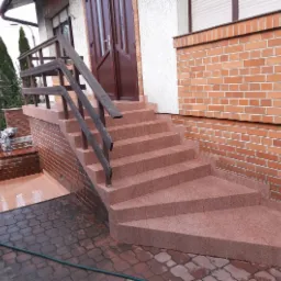 Zewnętrzne schody wejściowe do domu wyłożone płytkami w odcieniach czerwieni, z drewnianą balustradą i fragmentem muru z cegły klinkierowej.