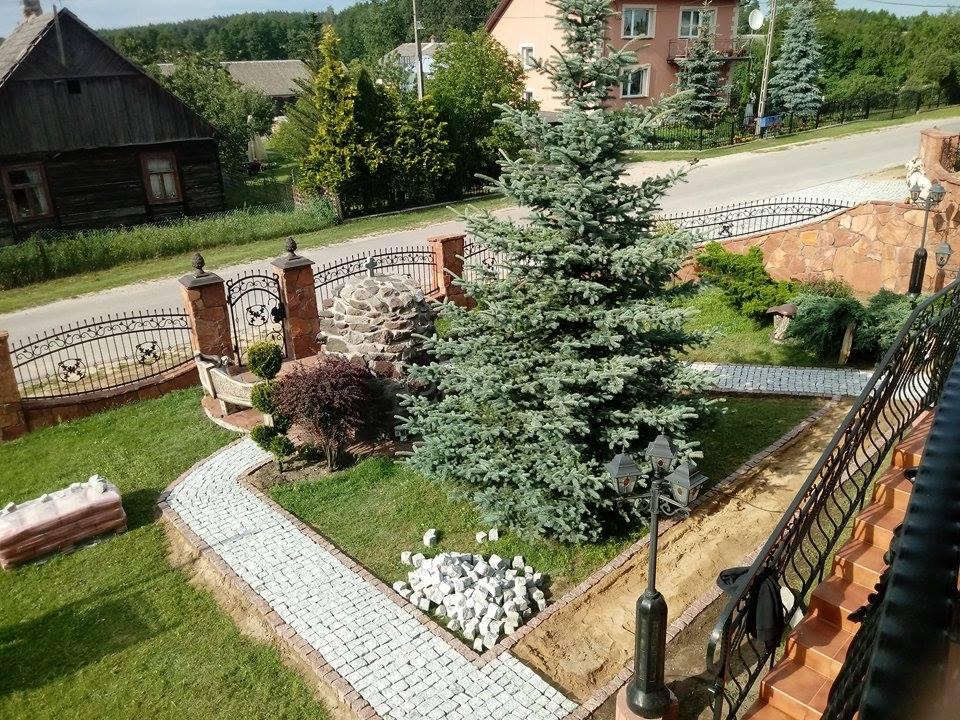 Fragment ogrodu z ścieżką z jasnej kostki granitowej w trakcie układania, widoczne przygotowane podłoże i stos kostki, w tle ozdobny murek z bramą i starodawnym domem.