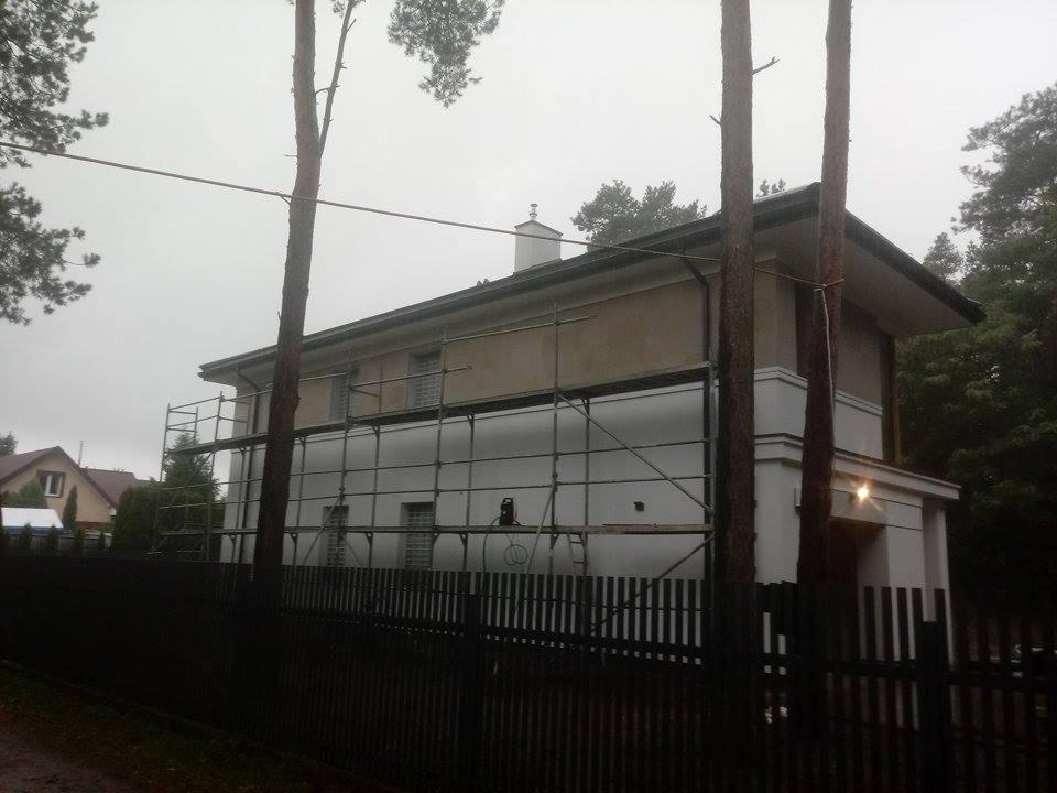 Dwupiętrowy dom w trakcie remontu elewacji, otoczony rusztowaniami, widok zza czarnego ogrodzenia w pochmurny dzień.