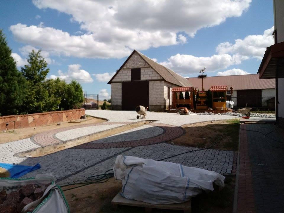 Ułożona kostka granitowa w kształcie łuków na podjeździe, widoczny budynek gospodarczy z brązową bramą, mieszarka do betonu i worki z kostką, błękitne niebo z chmurami.