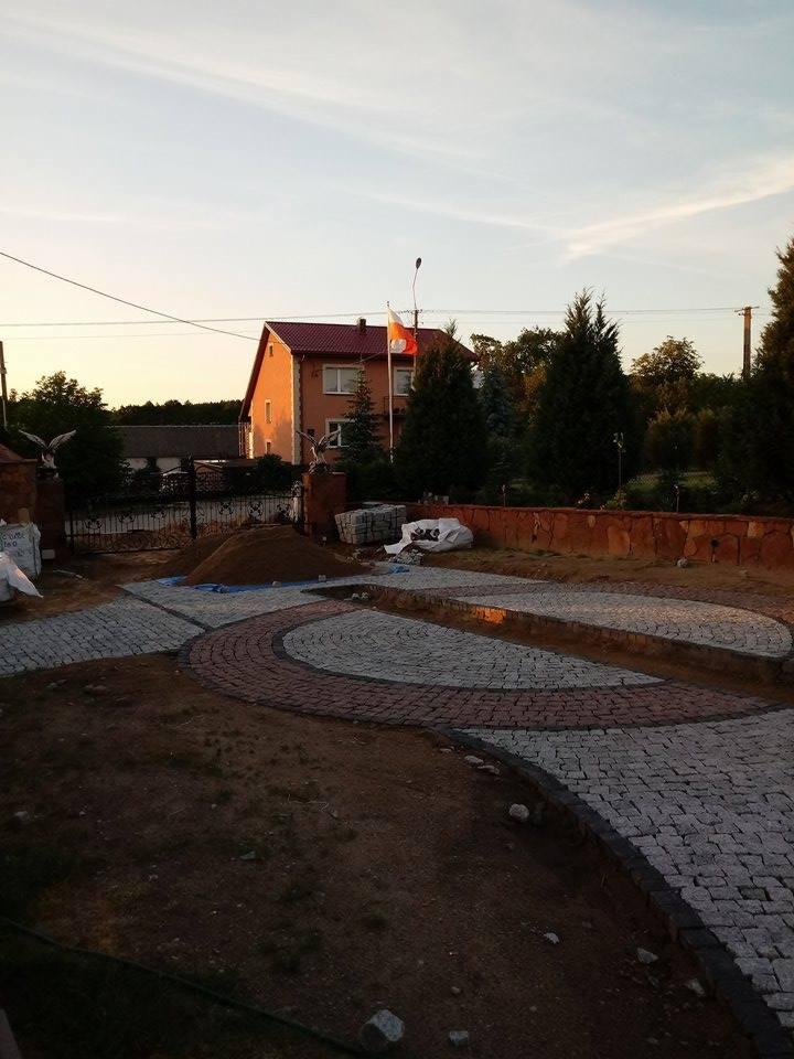 Kładzenie kostki granitowej w łukowaty wzór na posesji przed domem, widoczny kopiec piasku i przygotowane podłoże.