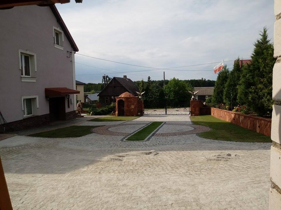 Ułożona kostka granitowa na podjeździe z elementami trawy i ozdobnymi obrzeżami, brama wjazdowa z kamiennymi słupkami zwieńczonymi orłami, w tle budynek mieszkalny i polska flaga.