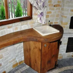 Rustykalna aranżacja wnętrza z drewnianym blatem, kamienną umywalką i murowanym piecem chlebowym w tle, widok z okna na zielone tuje.