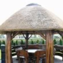 Altana ogrodowa z drewnianą konstrukcją i strzechą, otoczona trawnikiem i tujami, z drewnianym stołem i krzesłami wewnątrz.