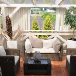 Zadaszony taras z brązowymi meblami rattanowymi, białymi poduszkami, dekoracyjnymi roślinami w białych donicach i zasłonami, widok z wnętrza na zewnątrz.