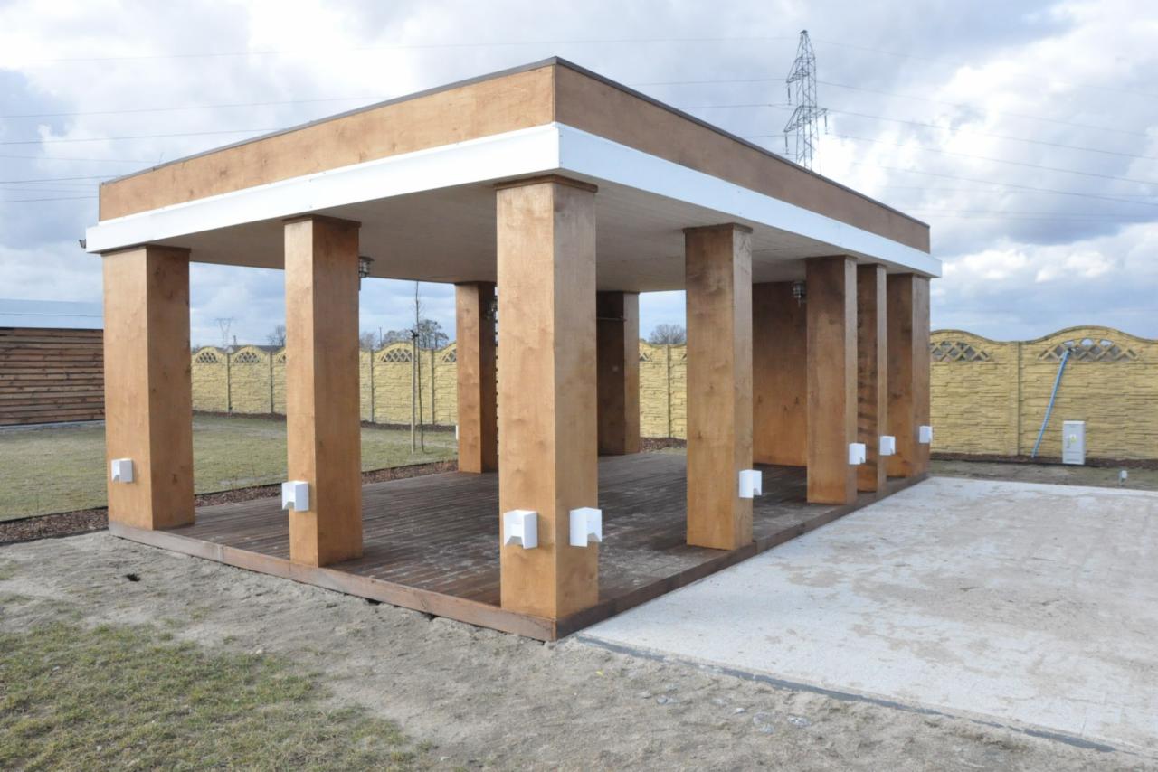 Nowoczesna altana z drewna, z płaskim dachem i białymi akcentami, oświetleniem na słupach, na drewnianym podeście, otoczona trawnikiem i ogrodzeniem.