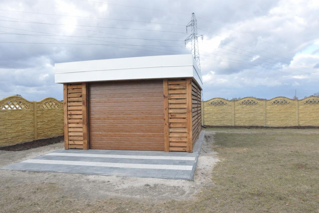 Mała, drewniana konstrukcja z brązową bramą rolowaną i białym dachem, otoczona ogrodzeniem z betonowych przęseł, umieszczona na wyłożonym szarymi płytami tarasie.