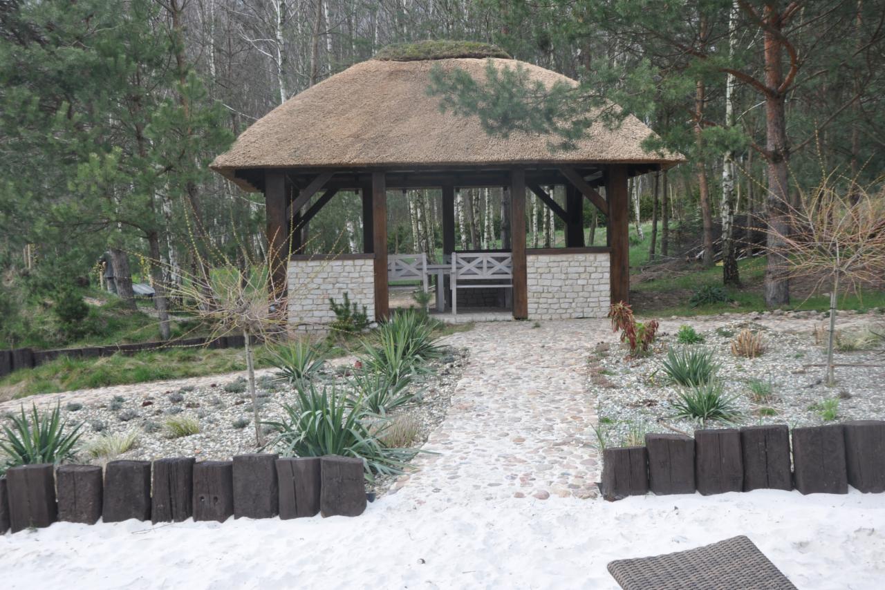 Zadaszona konstrukcja z drewnianymi słupami i kamiennymi ściankami, kryta strzechą, z białymi ławkami w środku, otoczona ogrodem z kamienną ścieżką i niską drewnianą palisadą.