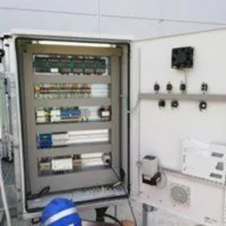 Otwarta szafa sterownicza z widocznymi elementami elektrycznymi, listwami zaciskowymi, bezpiecznikami i wentylatorem, obok leży niebieski kask.