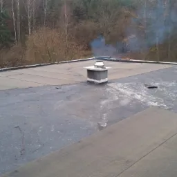 Czarny, płaski dach pokryty papą z widocznym kominem, butlą gazową z czerwonym wężem, puszką farby i narzędziami na tle drzew bez liści.