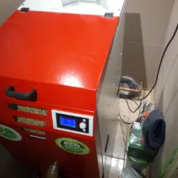 Czerwono-biały piec na pellet z widocznym panelem sterowania i zasobnikiem paliwa, w pomieszczeniu z instalacją hydrauliczną w tle.