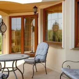 Zadaszony taras z oknami o drewnianych ramach, na zewnątrz dekoracyjny zegar, stolik i krzesła z niebiesko-białymi poduszkami w paski.