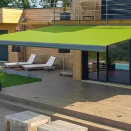 Zewnętrzna, rozłożona markiza w kolorze limonki nad tarasem z widokiem na basen i nowoczesny dom obłożony drewnem.