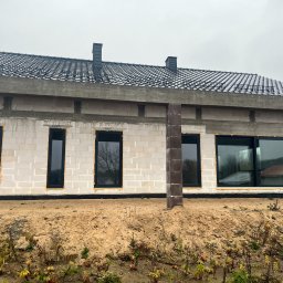 ECO-INSTAL GROUP SP. Z O.O. - Fasada nowo budowanego domu z białych bloczków, z zamontowanymi czarnymi oknami i drzwiami przesuwnymi, widoczną pianką izolacyjną. Ciemny dach z dachówkami i barierami śniegowymi, na tle...
