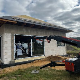 ECO-INSTAL GROUP SP. Z O.O. - Montaż dużych paneli szklanych w nowo powstającym budynku z białymi ramami, wspomagany przez mini-żuraw BG LIFT M250. Widoczne ściany z bloczków i konstrukcja dachu.