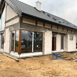 ECO-INSTAL GROUP SP. Z O.O. - Nowoczesny dom w stanie surowym zamkniętym, z jasnymi ścianami z bloczków, ciemnym dachem i dużymi oknami o brązowych ramach, w tym charakterystycznym, wysokim oknie trójkątnym. Piaszczyste...