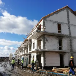 Budowa szeregu domów murowanych z betonu komórkowego, widoczne elementy konstrukcyjne dachu, rusztowania i pracownicy w odblaskowych kamizelkach na błotnistym terenie.