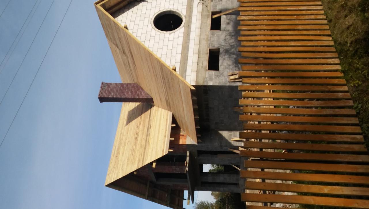 Wznoszony budynek z białego bloczka z okrągłym oknem, widoczne drewniane szalunki i rusztowanie, komin z cegły. Ujęcie z dołu pod kątem.