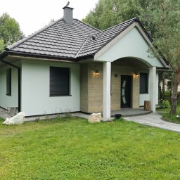 Paweł Potoczny - Nowoczesny dom jednorodzinny z szarą dachówką i elementami drewnianej elewacji, otoczony zielonym trawnikiem z ozdobnymi kamieniami. Widoczne rolety zewnętrzne.