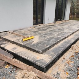 Paweł Potoczny - Nowoczesny taras z szarej kostki brukowej w trakcie budowy, z widoczną szczotką i piaskiem, przylegający do jasnego budynku z dużymi oknami.