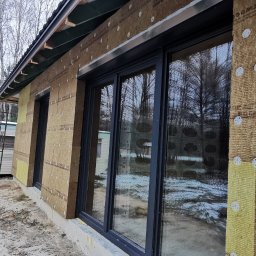 Paweł Potoczny - Ocieplanie ścian zewnętrznych domu wełną mineralną, widoczne mocowania i okna. Prace w toku, teren budowy. Częściowo ocieplone ściany budynku z nowymi oknami.