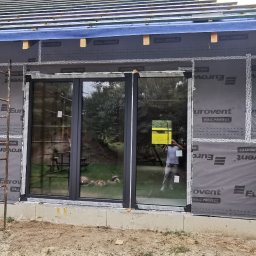 Paweł Potoczny - Montaż okien w domu szkieletowym, widoczne rusztowania i membrana Eurovent. Prace budowlane w toku, z zewnątrz widok na zieleń.