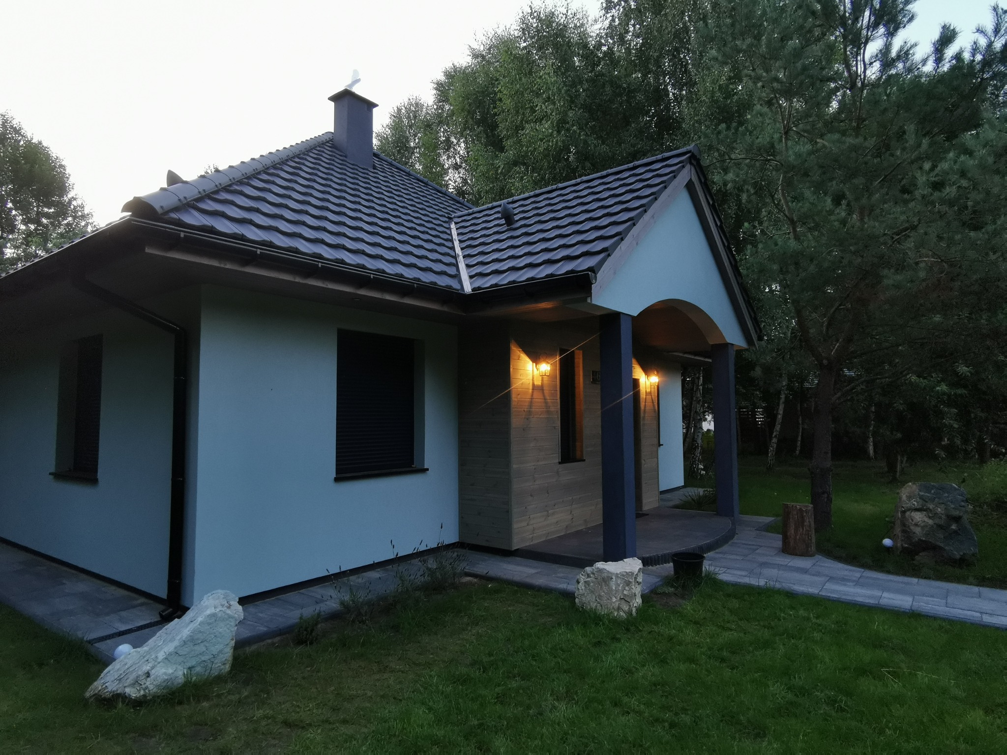 Nowoczesny dom z szarą dachówką i elementami drewna, oświetlony ciepłym światłem. Widoczne kamienne dekoracje w ogrodzie i ścieżka z kostki.