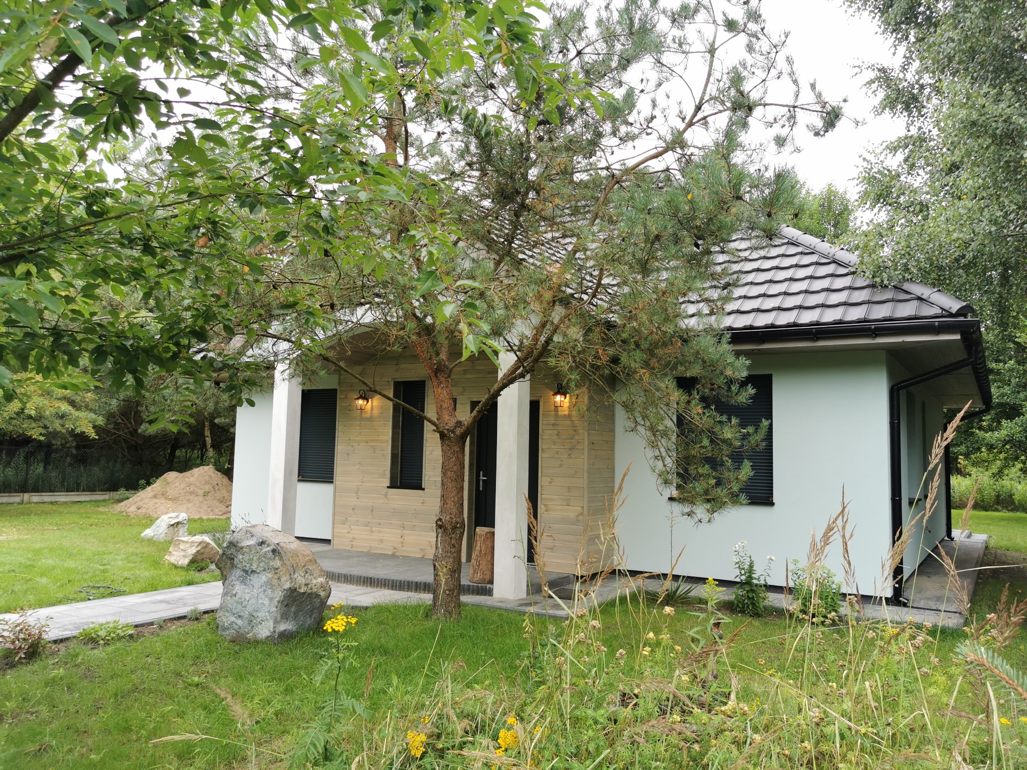 Parterowy dom z drewnianą elewacją częściowo zasłonięty drzewem. Widoczne lampy przy drzwiach i kamień dekoracyjny w ogrodzie. Ujęcie z poziomu gruntu.