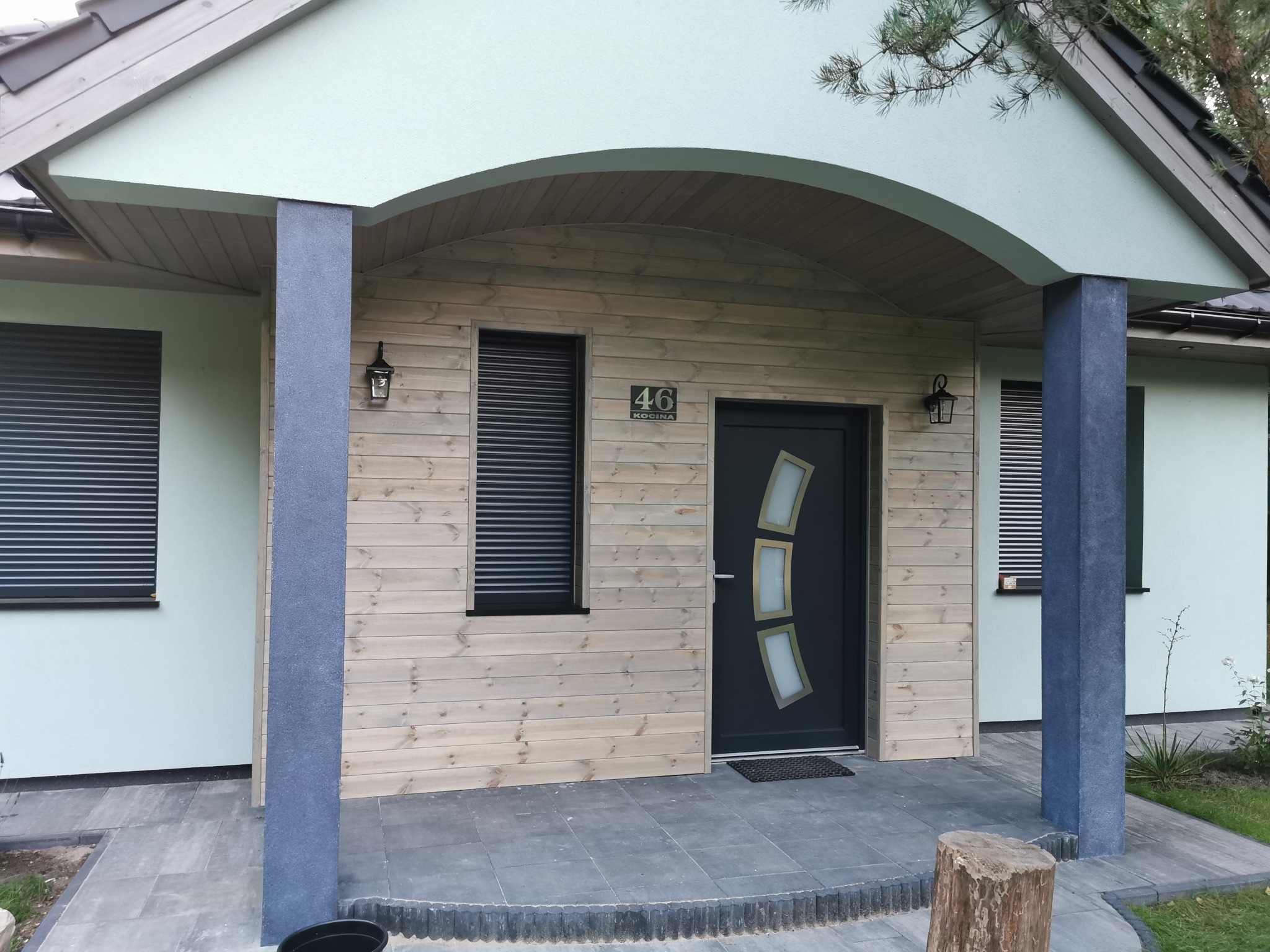Elewacja domu z jasnego drewna z numerem 46, czarne rolety w oknach, ciemne drzwi wejściowe z ozdobnymi szybami i kolumny w kolorze grafitowym.