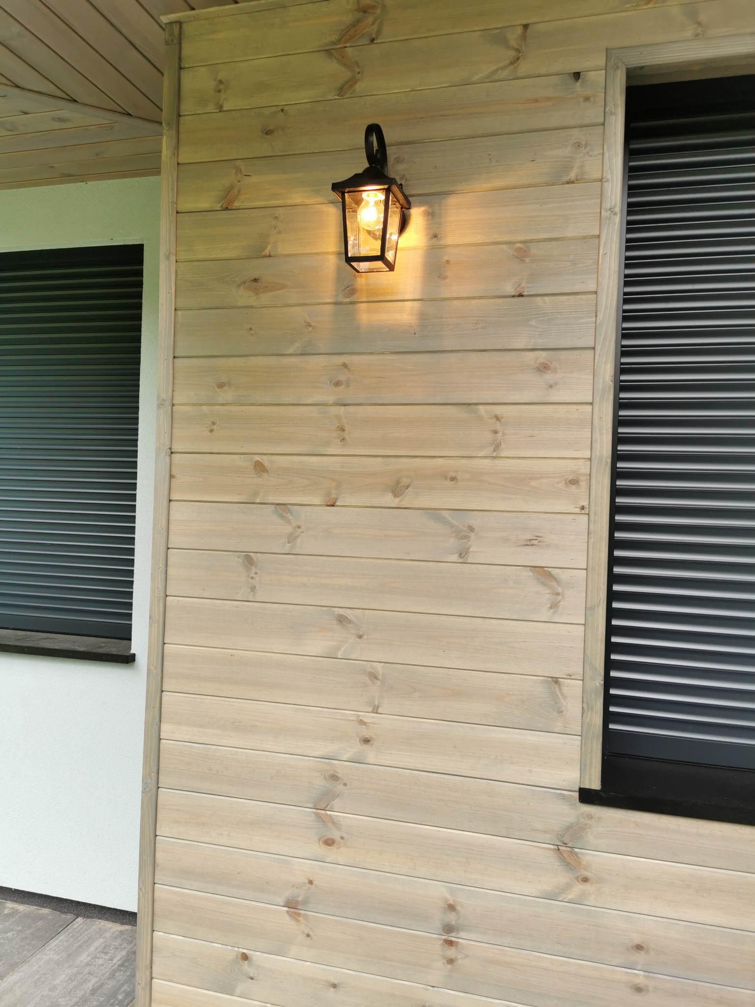 Drewniana elewacja domu z oknem i lampą w Kocinie. Jasne deski ułożone poziomo, czarne rolety i ozdobna lampa zewnętrzna.
