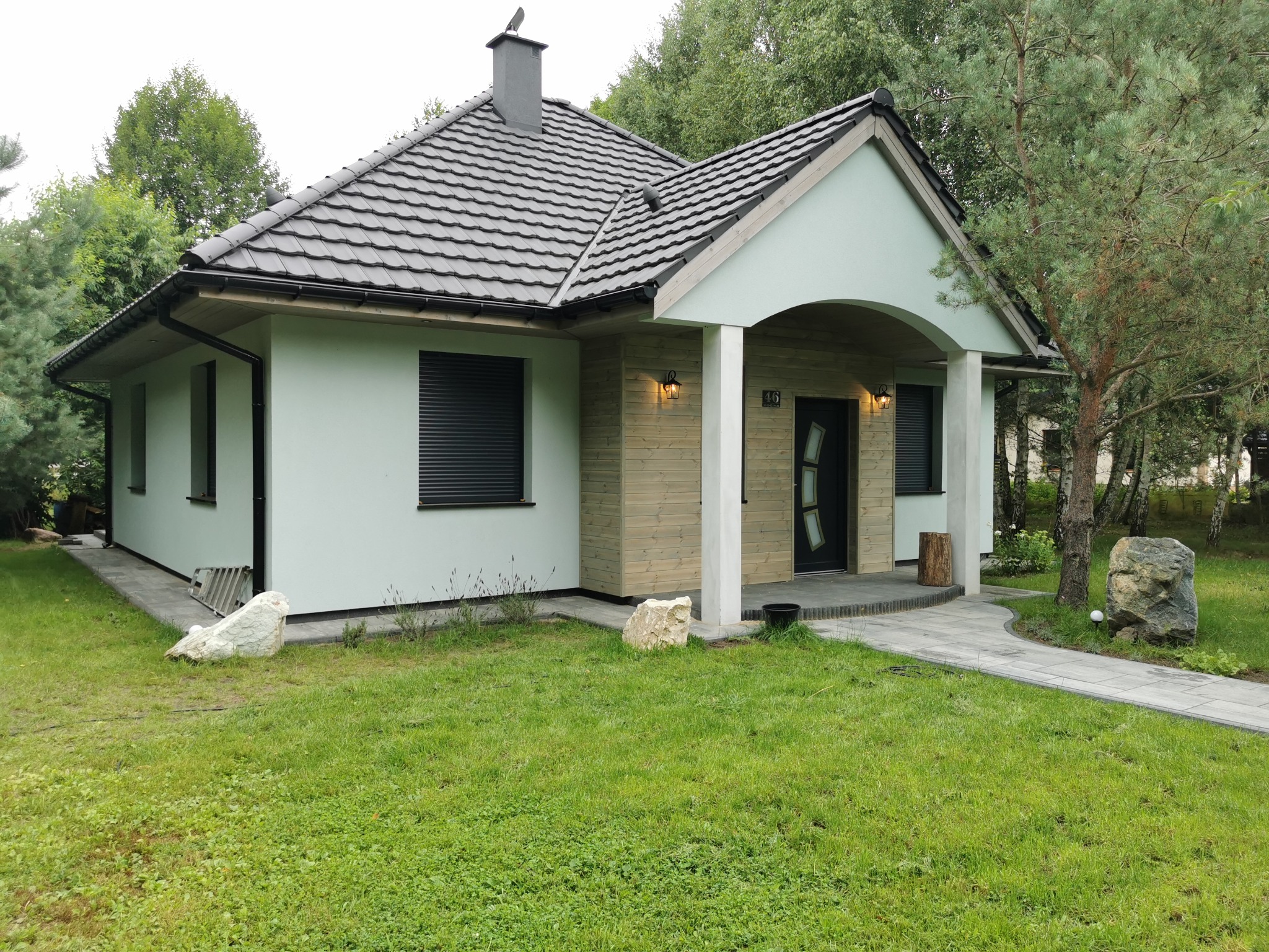Nowoczesny dom jednorodzinny z szarą dachówką i elementami drewnianej elewacji, otoczony zielonym trawnikiem z ozdobnymi kamieniami. Widoczne rolety zewnętrzne.