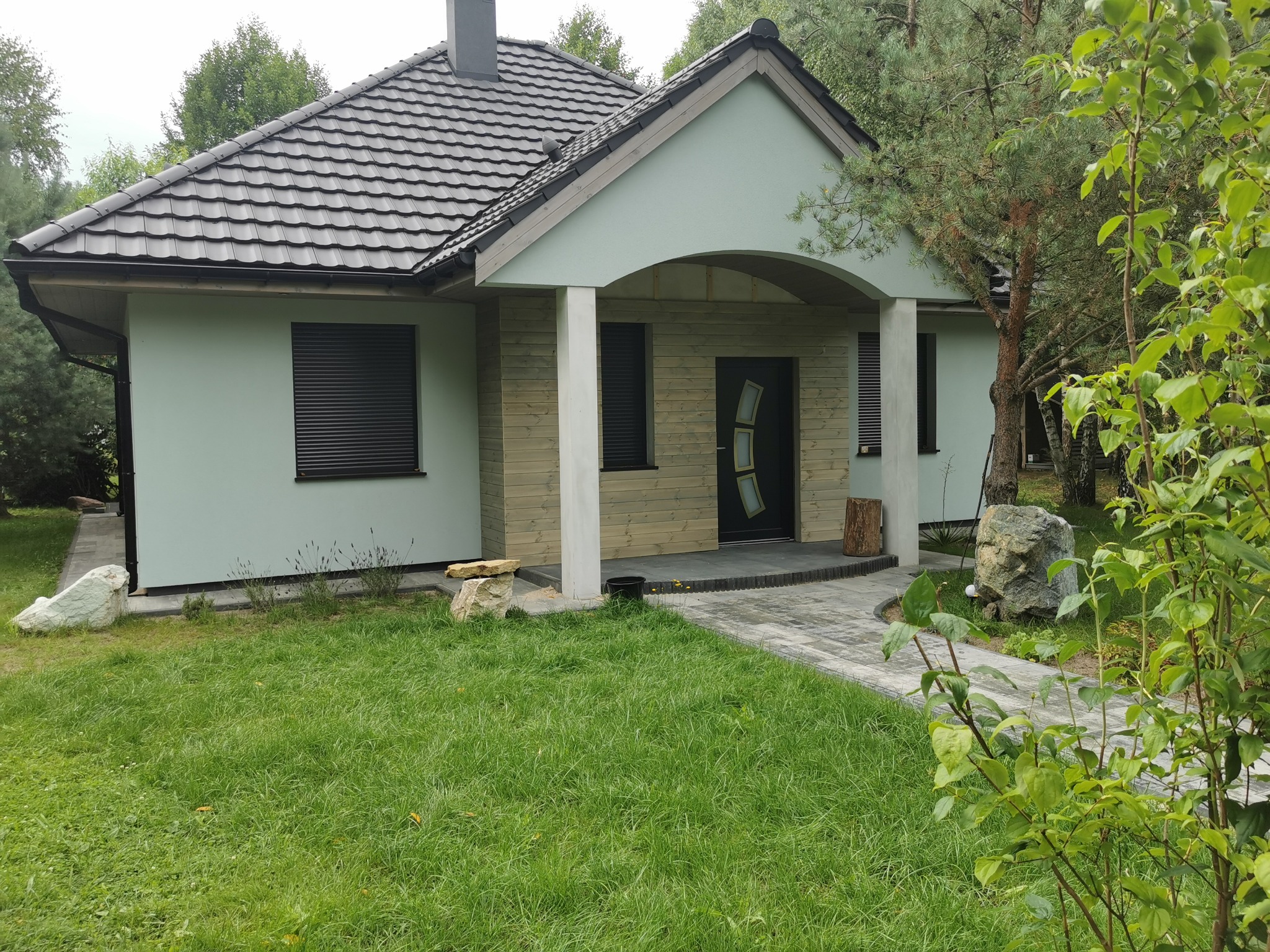 Gotowy dom szkieletowy z zieloną elewacją i ciemną dachówką, otoczony zielenią. Widoczny fragment chodnika z kostki brukowej i elementy dekoracyjne z kamienia.