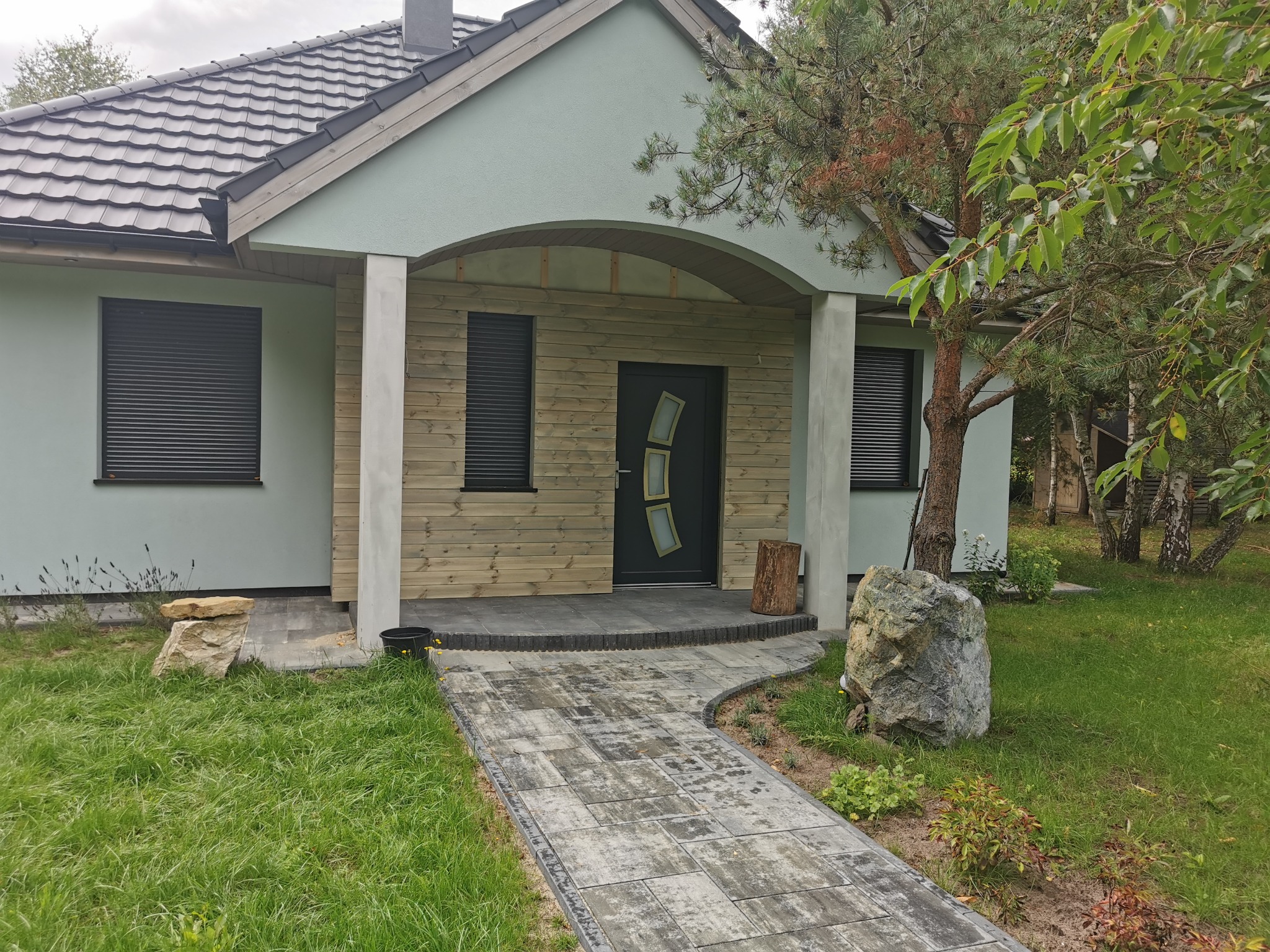 Gotowy dom szkieletowy z podjazdem z kostki, widok z zewnątrz. Elementy drewniane i jasna elewacja. Uwagę przykuwają duże kamienie wkomponowane w zieleń.