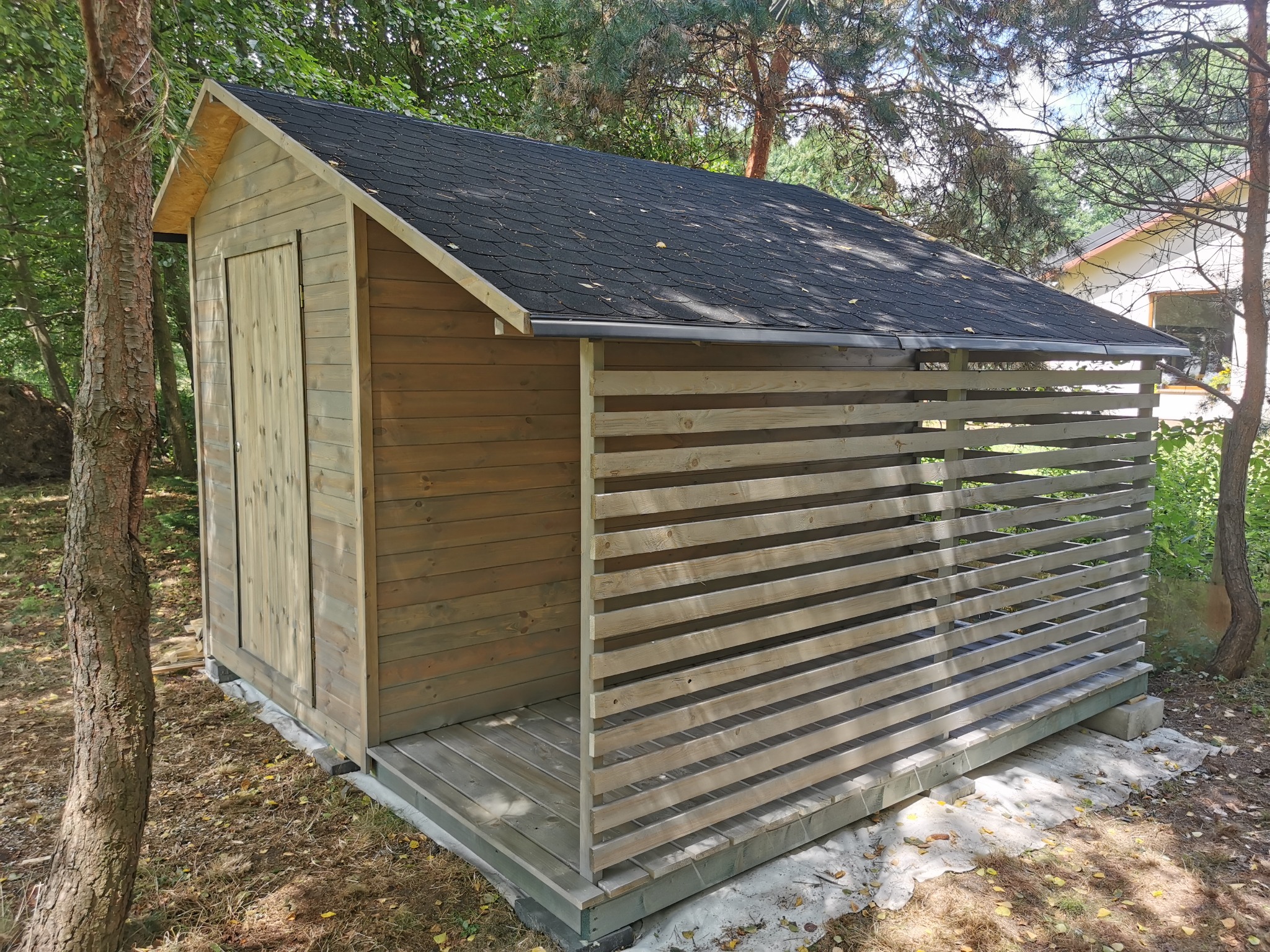 Drewniana altana z zamykanymi drzwiami i ażurową ścianą boczną, kryta papą, usytuowana w ogrodzie, widziana z perspektywy lekko z góry.