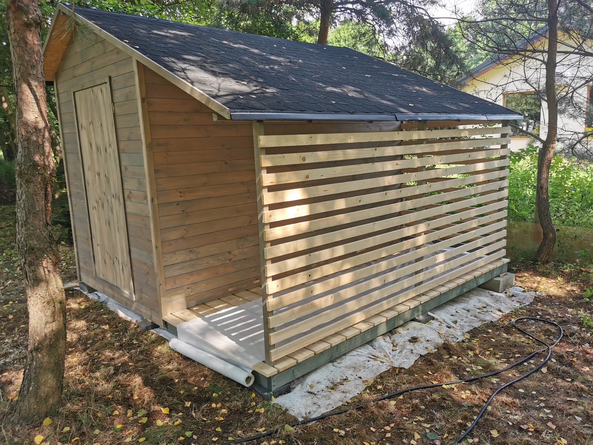 Drewniany domek narzędziowy z zadaszeniem i ażurową ścianą z desek, umieszczony na betonowych bloczkach. Widoczne drzewa w tle, czarny wąż ogrodowy na ziemi.