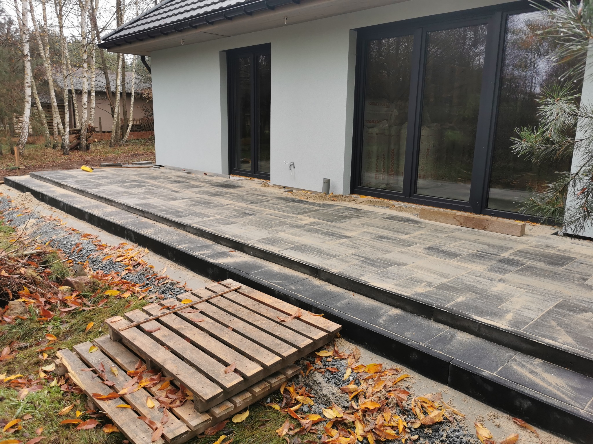 Nowoczesny taras z szarych płyt, z niskimi schodami i paletą obok. Widoczne duże okna domu i jesienne liście. Taras w trakcie budowy.