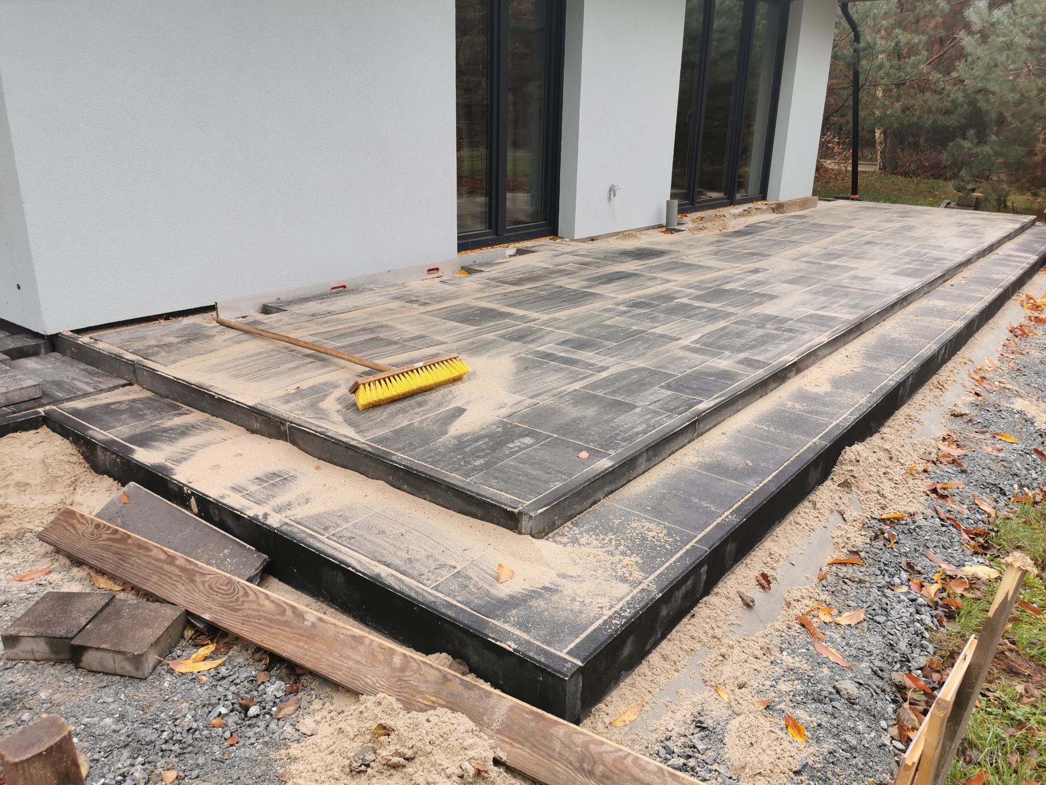 Nowoczesny taras z szarej kostki brukowej w trakcie budowy, z widoczną szczotką i piaskiem, przylegający do jasnego budynku z dużymi oknami.
