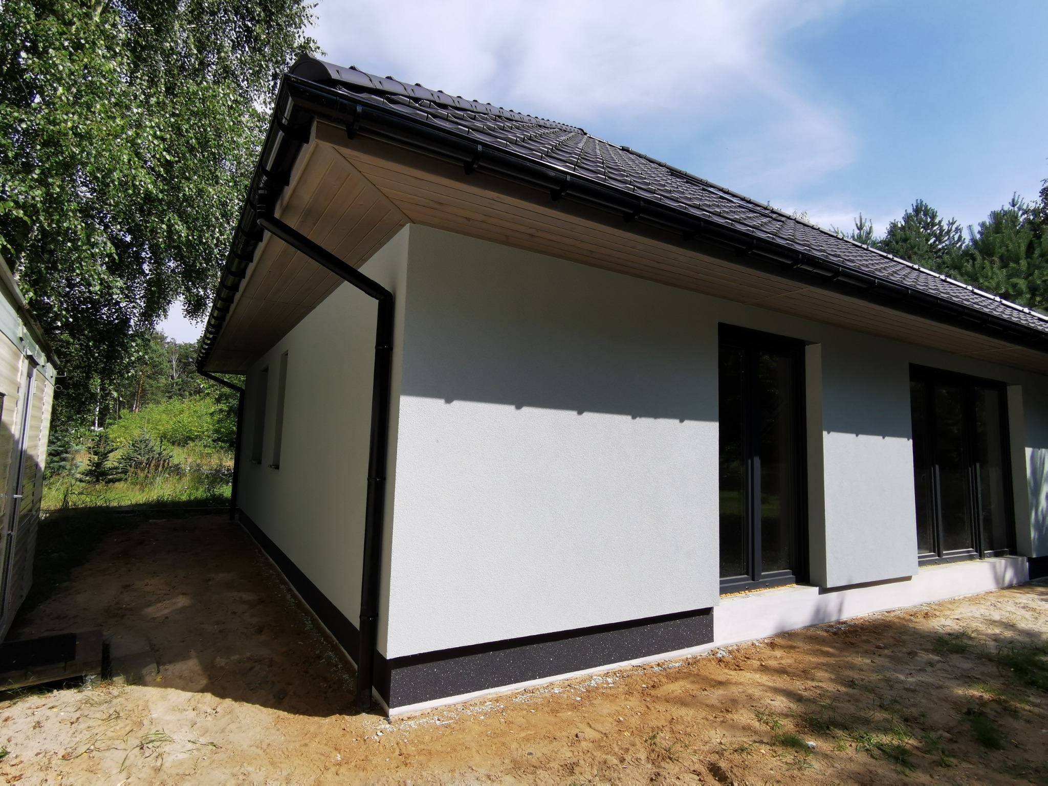 Nowoczesny, parterowy dom z szarą elewacją, czarną podmurówką i dachem z ciemnej dachówki. Widok z boku, z drzewami w tle i fragmentem budynku gospodarczego po lewej.