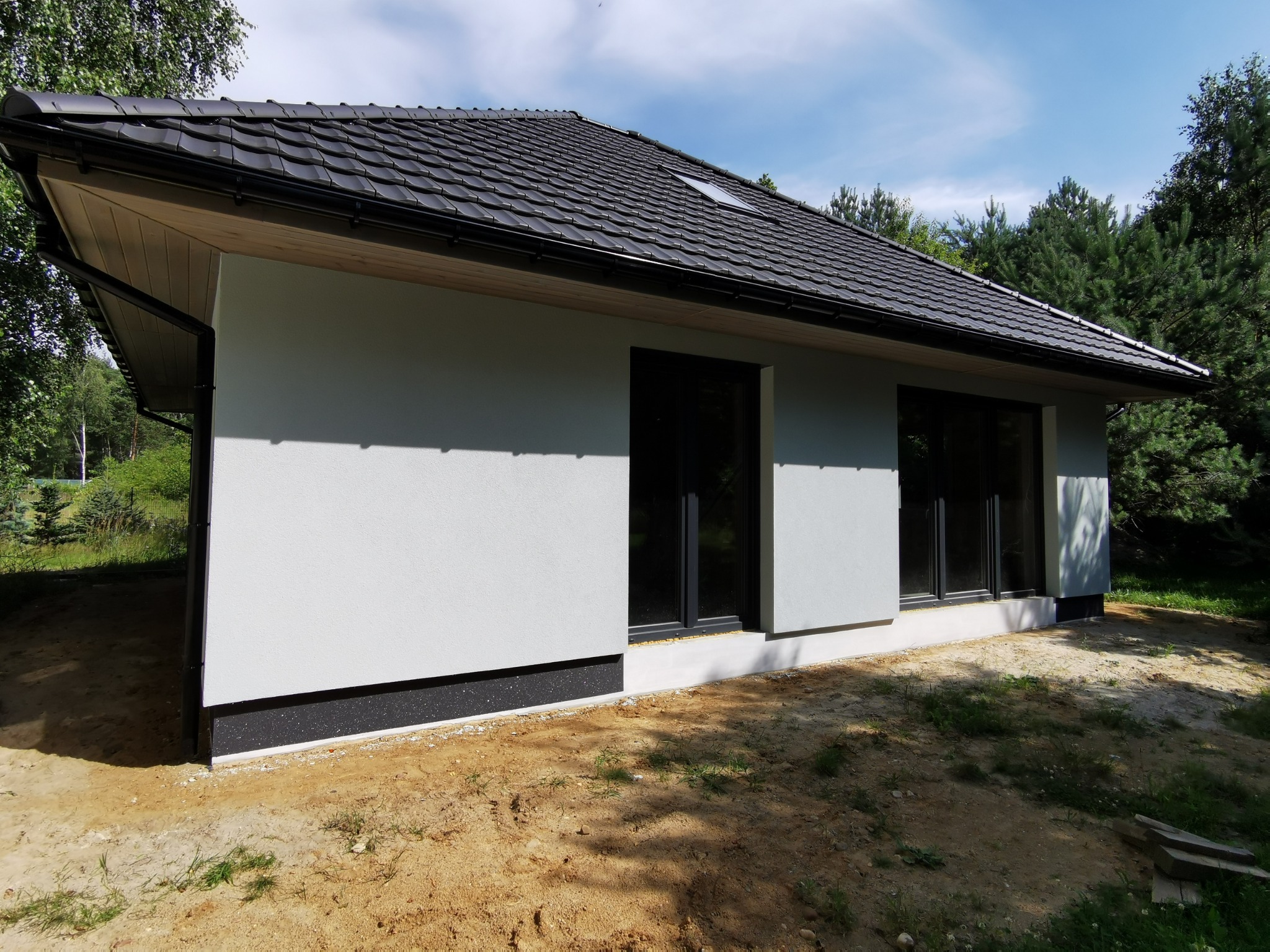 Niski, nowoczesny dom z szarą elewacją, czarnym dachem z dachówki i dwoma oknami. Widoczne detale wykończenia oraz fragment ogrodu. Ujęcie z boku budynku.