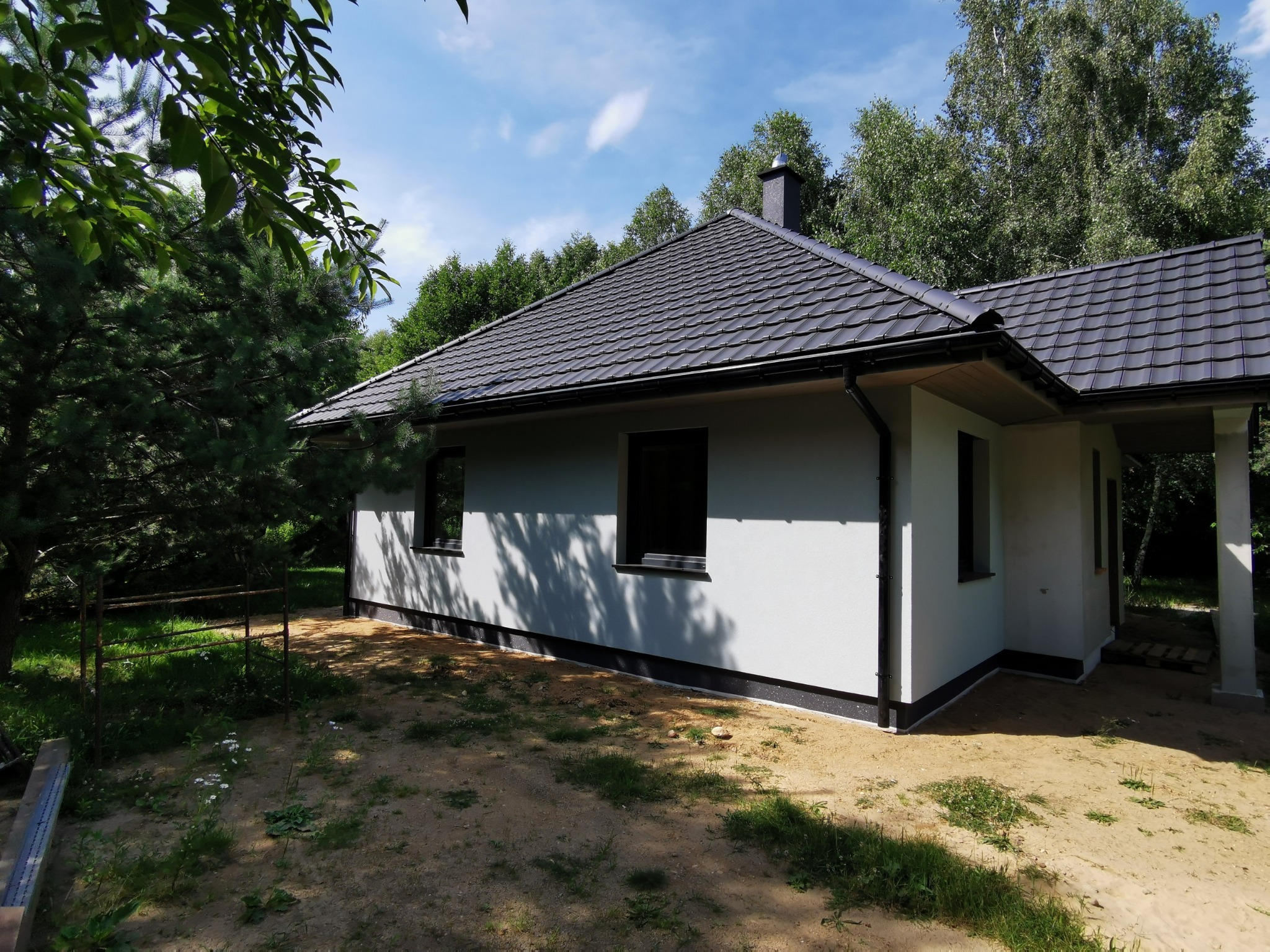 Nowoczesny dom z ciemną dachówką i jasną elewacją, otoczony zielenią. Widoczne okna z czarnymi ramami i fragment konstrukcji tarasu.