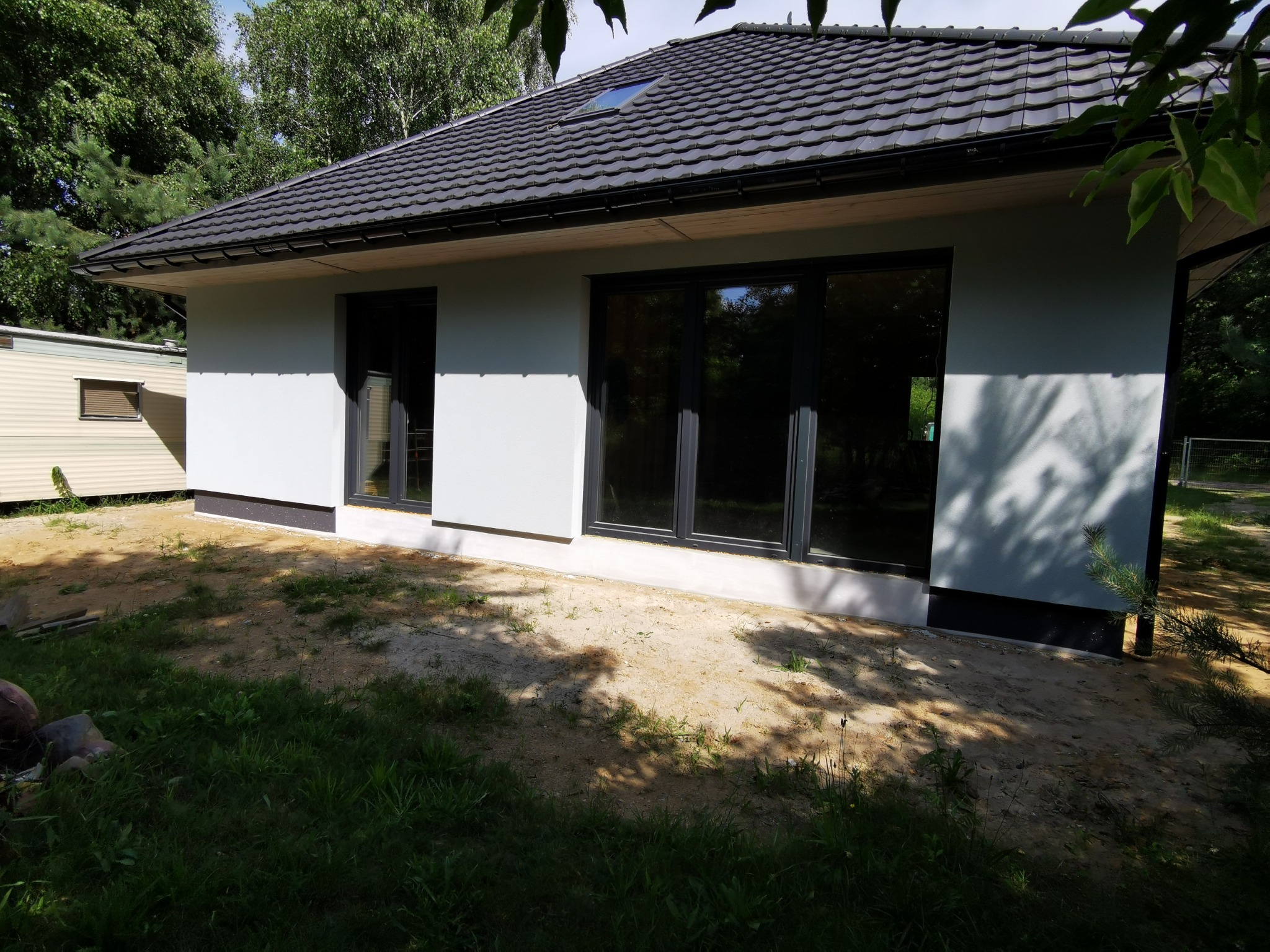 Parterowy dom z szarą elewacją i ciemną dachówką, duże okna z czarnymi ramami. Widoczne drzewa i trawa. Obok przyczepa kempingowa. Ujęcie z zewnątrz.