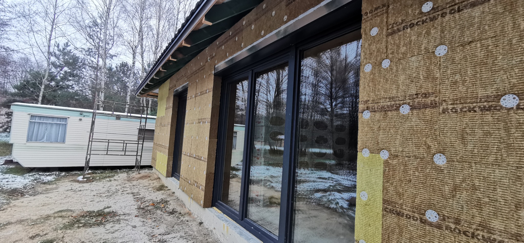 Ocieplanie ścian zewnętrznych domu wełną mineralną, widoczne mocowania i okna. Prace w toku, teren budowy. Częściowo ocieplone ściany budynku z nowymi oknami.