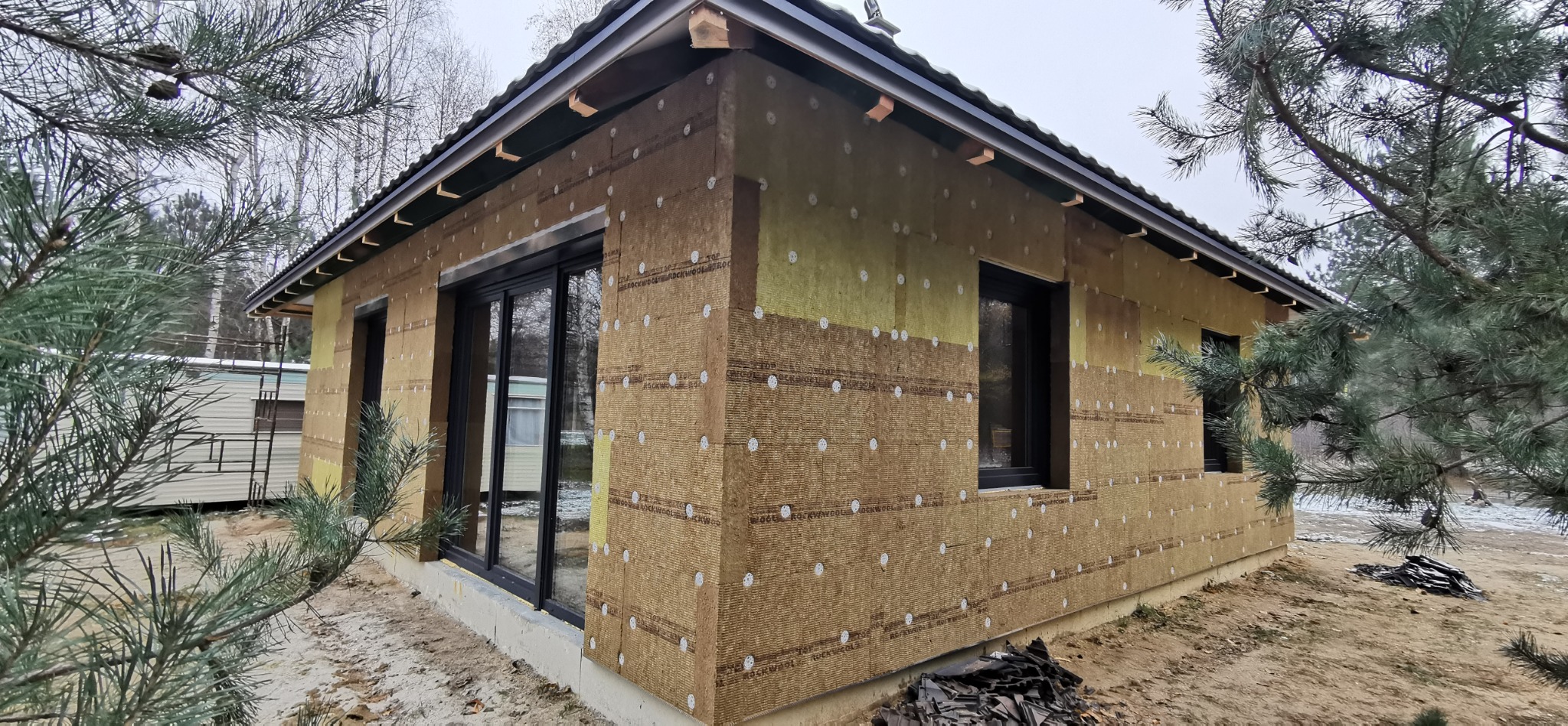 Dom szkieletowy w trakcie ocieplania, widoczne płyty izolacyjne z wełny mineralnej z białymi kołkami mocującymi, czarne okna i fragment dachu, na zewnątrz drzewa iglaste.