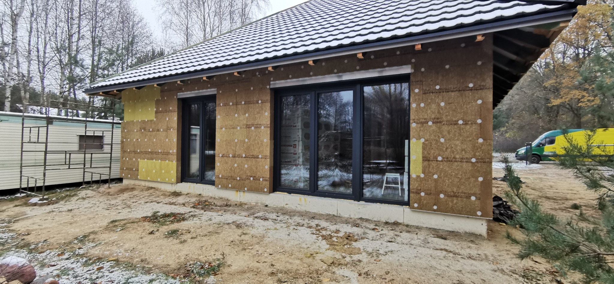 Budowa domu szkieletowego: ściany z izolacją, czarne okna, biała dachówka. Na zewnątrz zaparkowany zielono-żółty bus.