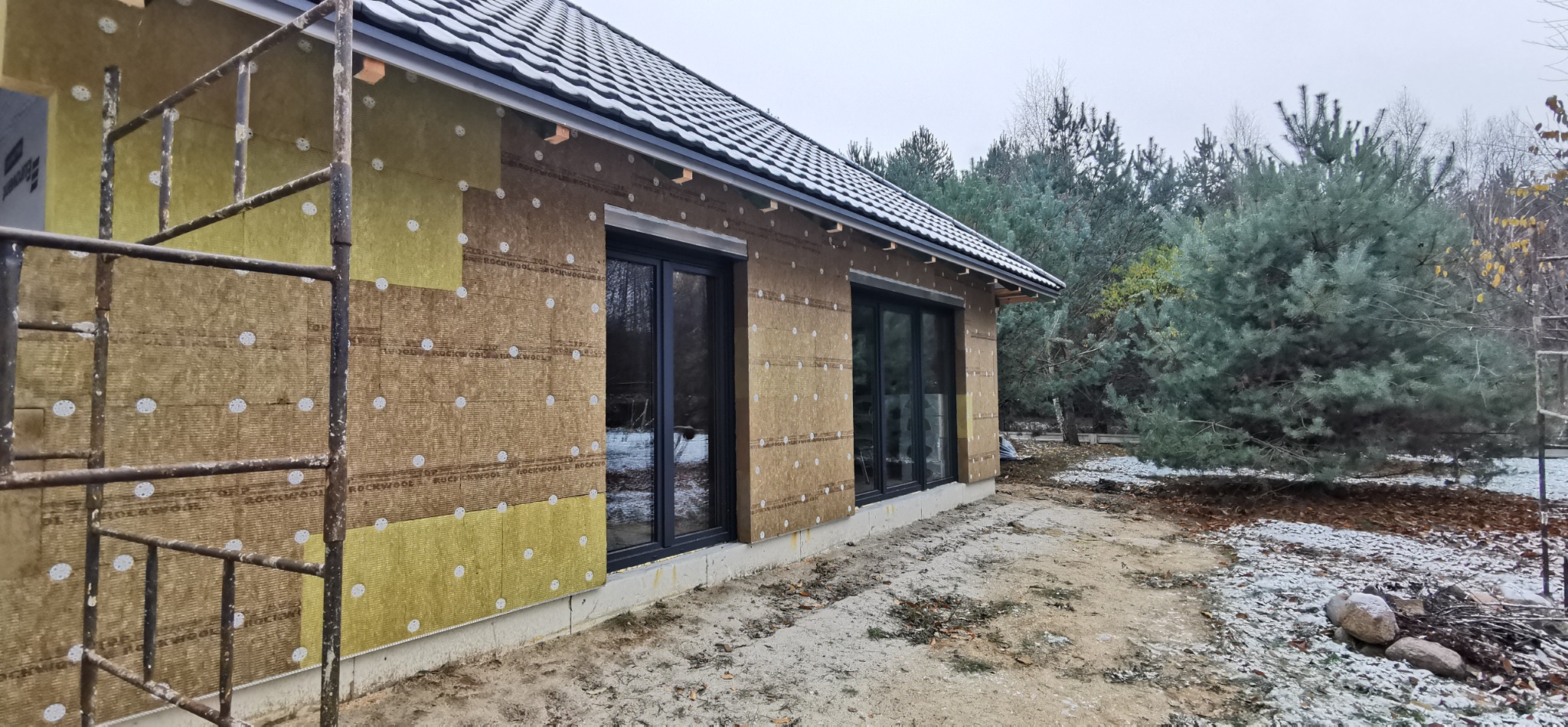 Dom szkieletowy w trakcie budowy: ściany z izolacją z wełny mineralnej, widoczne rusztowanie. Na dachu leży śnieg. W tle drzewa.
