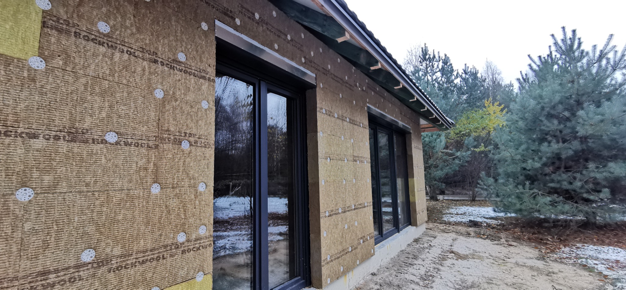 Ściana domu w trakcie ocieplania wełną mineralną Rockwool, widoczne okna z czarnymi ramami i elementy mocujące izolację. W tle drzewa.