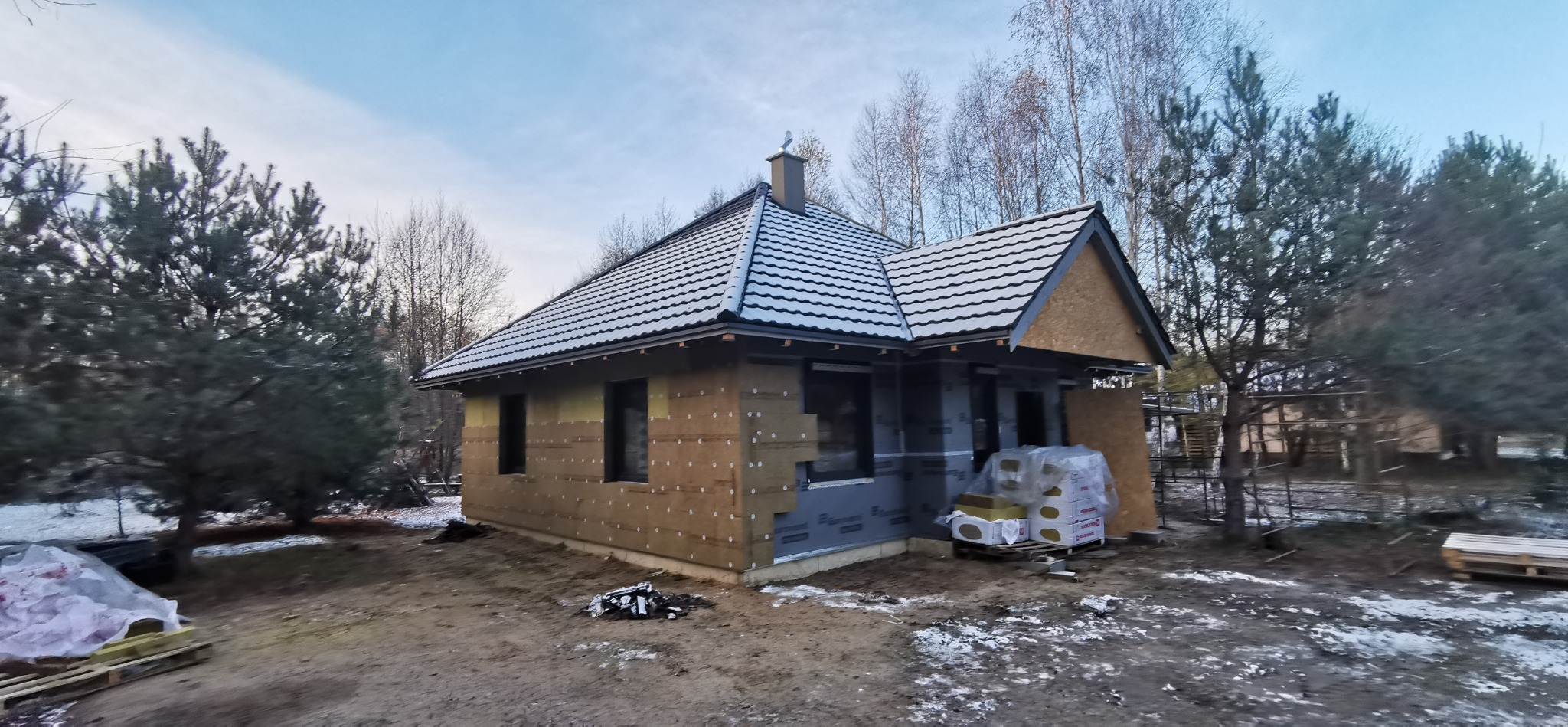 Niedokończony dom szkieletowy z widocznym ociepleniem i membraną, na tle drzew i zimowego krajobrazu. Dach pokryty dachówką.