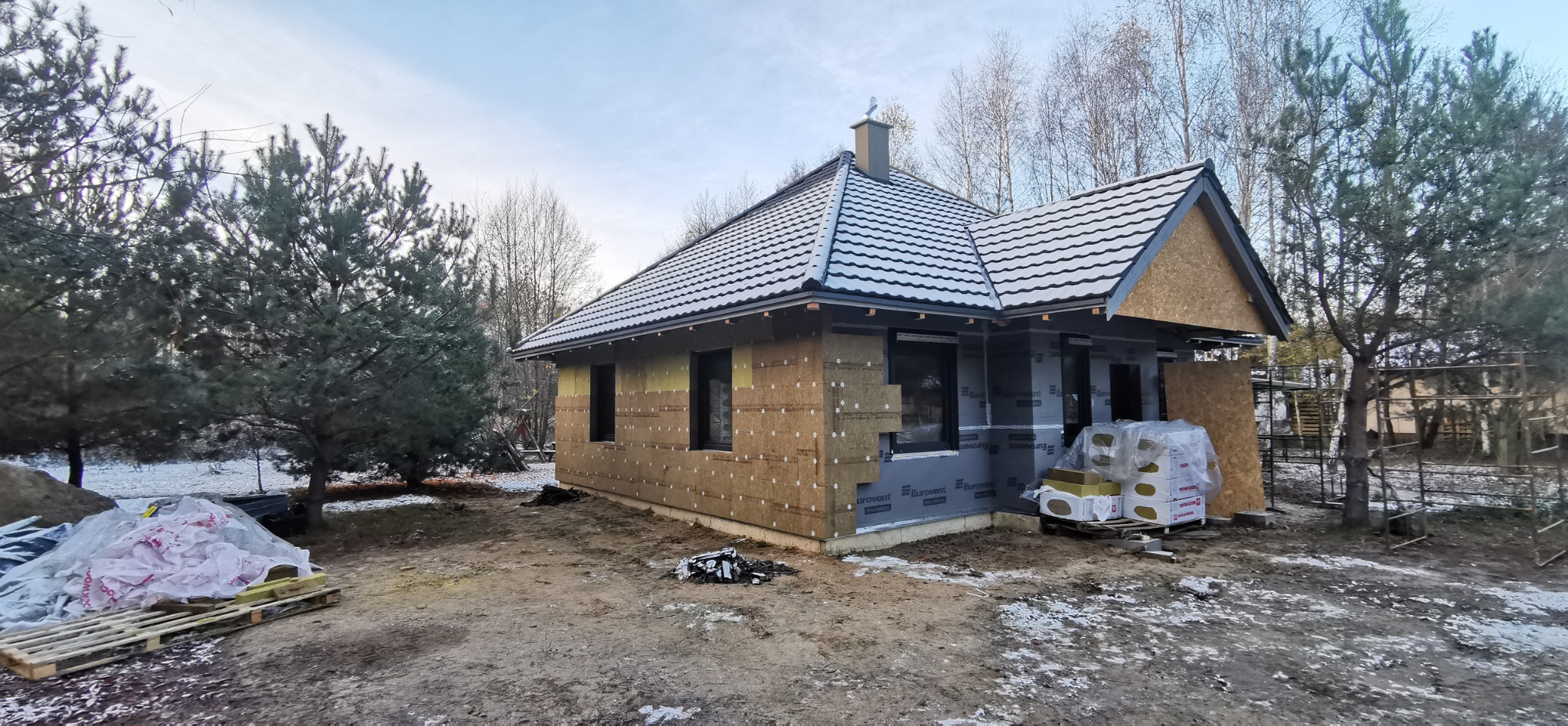 Budowa domu szkieletowego w trakcie: widoczne ocieplenie ścian wełną mineralną i membrana. Część elewacji wykończona. Na dachu dachówka. Zimowy krajobraz.