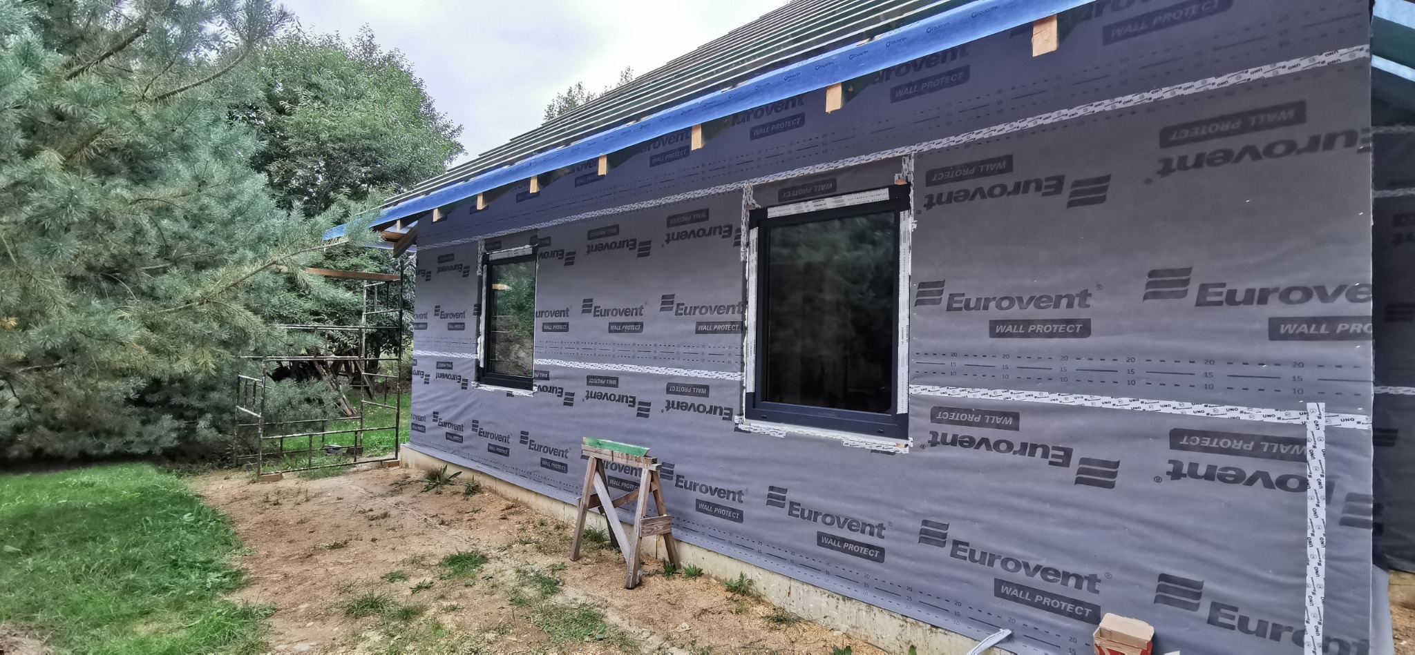 Dom szkieletowy w trakcie budowy: widoczne okna, folia izolacyjna Eurovent i elementy konstrukcji dachu. W tle zieleń drzew i krzewów, drabina. Ujęcie z zewnątrz.