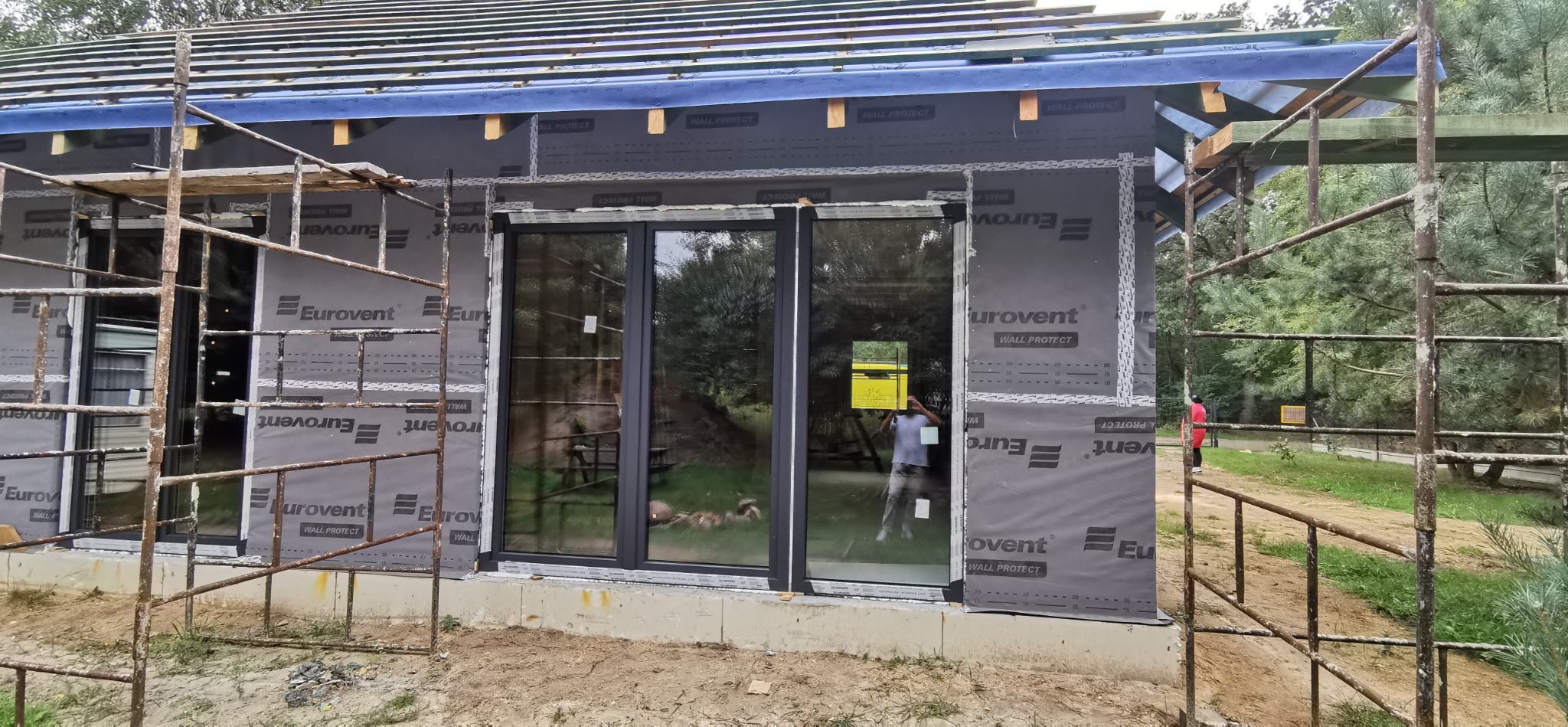 Montaż okien w domu szkieletowym, widoczne rusztowania i membrana Eurovent. Prace budowlane w toku, z zewnątrz widok na zieleń.